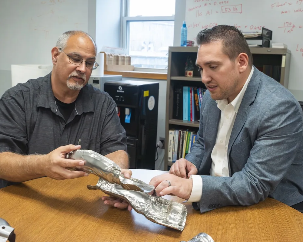 Two Soundwich team members looking at a heat shield on an office table, in discussion Two team members looking at a heat shield on an office table, discussing Soundwich's capabilities